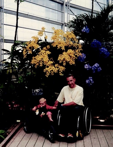 Afbeelding van vader in een rolstoel met dochter in een buggy aan het poseren in een plantentuin